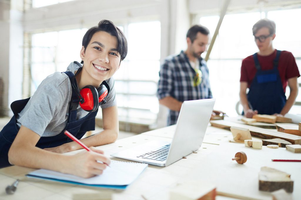 Excited lady enjoying work in carpentry Cheerful excited young lady with ear protectors on neck leaning on table and smiling at camera while enjoying work in carpentry